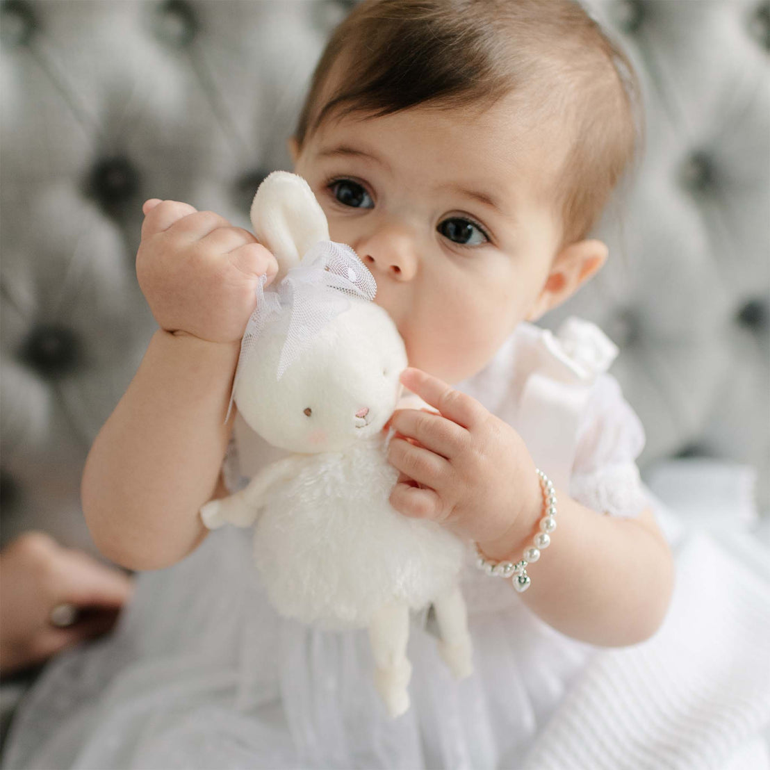 Baby holding a Stella Roly Poly Bunny against a grey cushioned sofa. Details of the bunny include a Stella lace Bow around its ears. Perfect for unique baby gifts.