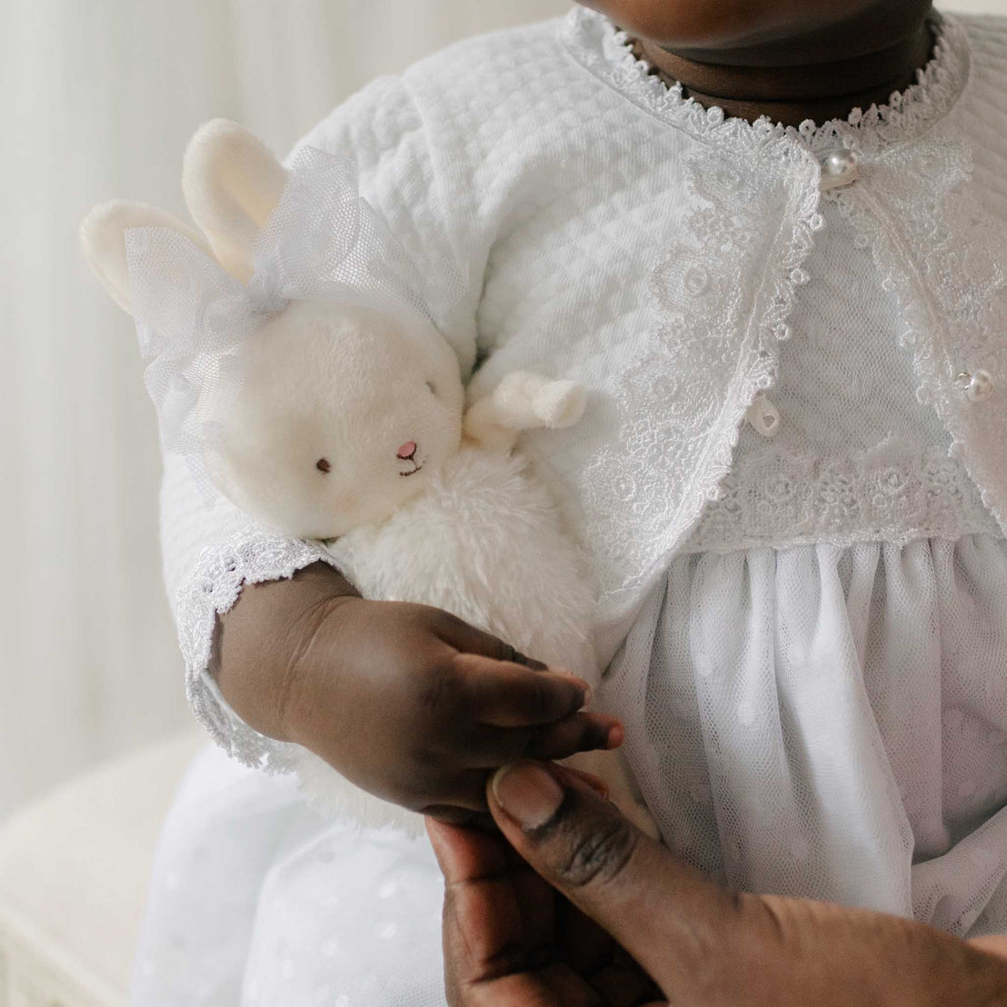 Close up of a baby holding the Stella Roly Poly Bunny against a soft white background. The plush bunny includes a Stella lace bow tied around its ears. Perfect for unique baby gifts.