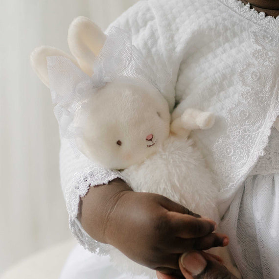 Close up of a baby holding the Stella Roly Poly Bunny against a soft white background. The plush bunny includes a Stella lace bow tied around its ears. Perfect for unique baby gifts.