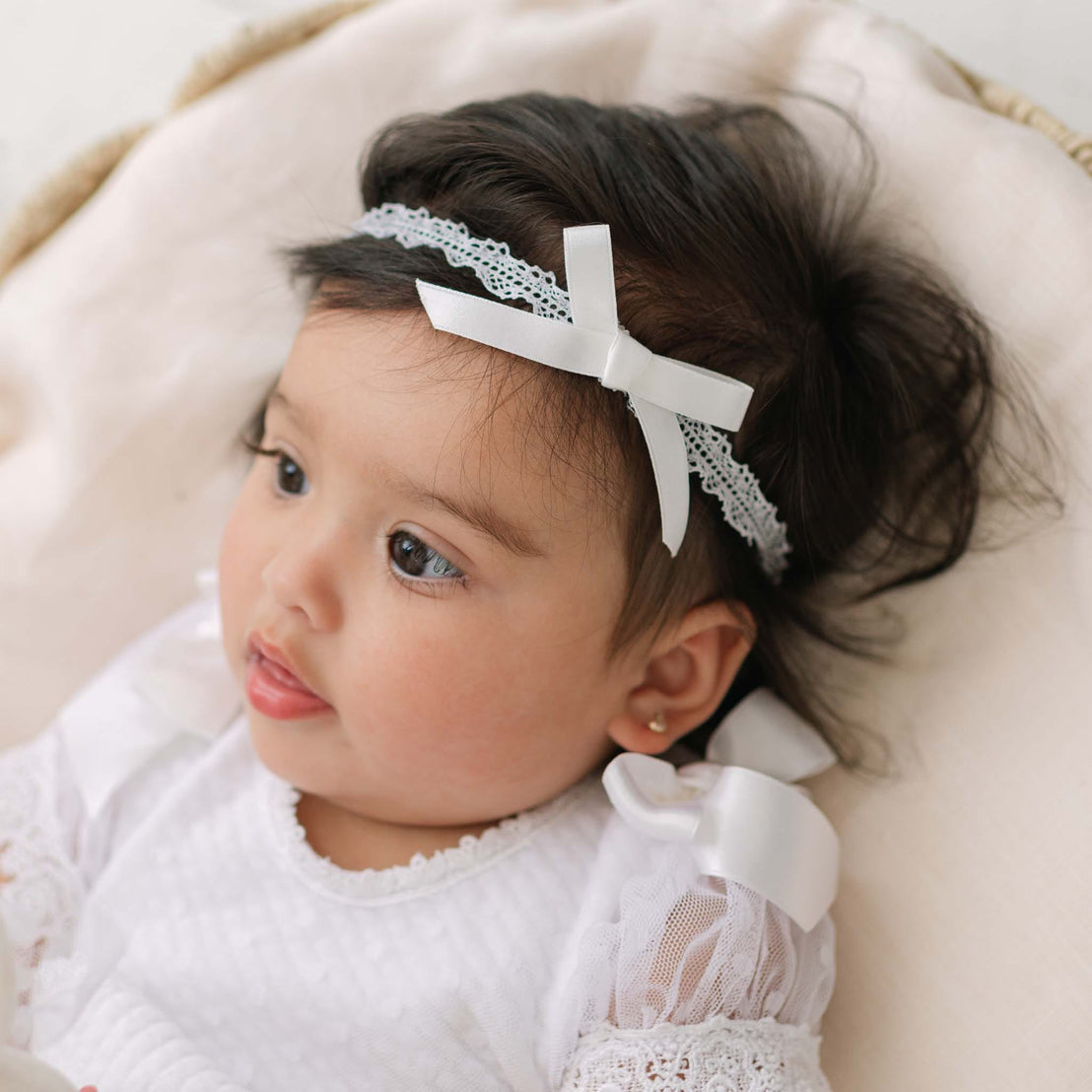 Baby laying in a wicker bassinet and wearing the Stella Silk Ribbon Headband and matching Dress. Details of the headband include white Venice lace with a silk ribbon bow.