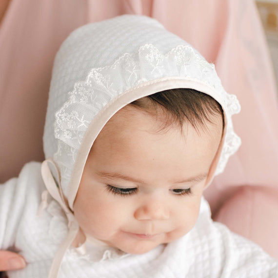 Newborn baby girl wearing the Tessa Quilt Bonnet gazes down.  The angle showcases the bonnet's edge details in pink champagne silk and light ivory floral lace.