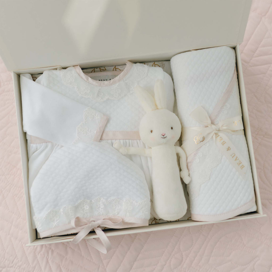 The Tessa Newborn Gift Set, currently available with a 10% discount, includes a neatly arranged collection featuring a white quilted dress made from soft pima cotton, complemented by a long-sleeve top, matching pants, an adorable cuddly toy rabbit, and a rolled-up blanket tied with a ribbon. These items are beautifully displayed against a pale pink quilted background.