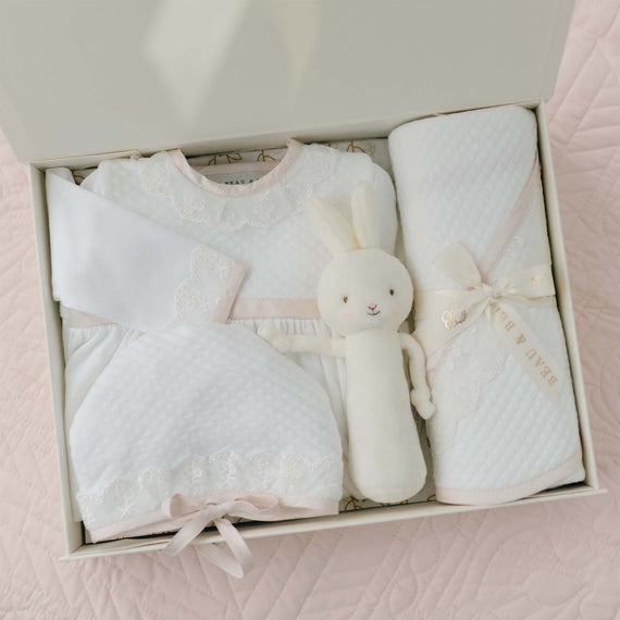 The Tessa Newborn Gift Set, currently available with a 10% discount, includes a neatly arranged collection featuring a white quilted dress made from soft pima cotton, complemented by a long-sleeve top, matching pants, an adorable cuddly toy rabbit, and a rolled-up blanket tied with a ribbon. These items are beautifully displayed against a pale pink quilted background.