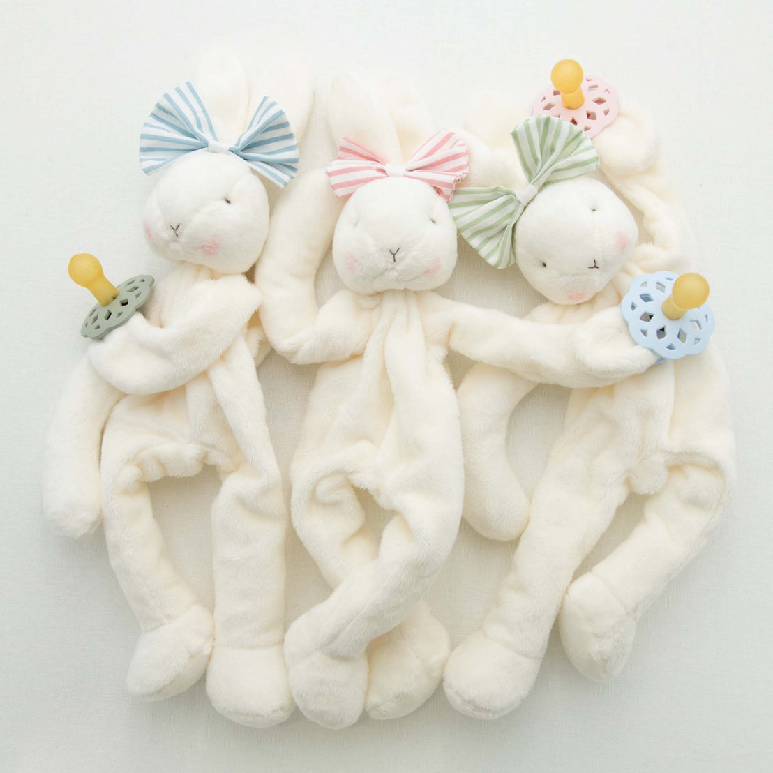 Three Thea Bunny Buddy toys, each wearing a miniature version of the Thea Bow Headband in blue, pink, green and holding a matching pacifier, lying on a white background.