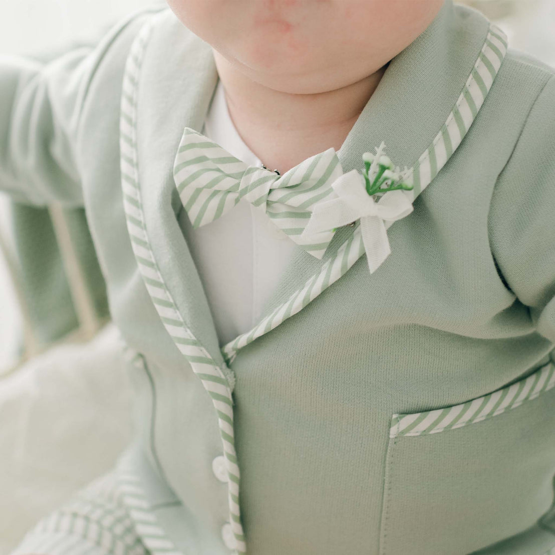 A close-up of a baby dressed in the Theodore Bow Tie & Boutonniere, featuring a pastel green outfit with white striped trim. The ensemble is complete with a matching handmade bow tie and a small decorative boutonniere with locking pin pinned to the lapel. The background is softly blurred.