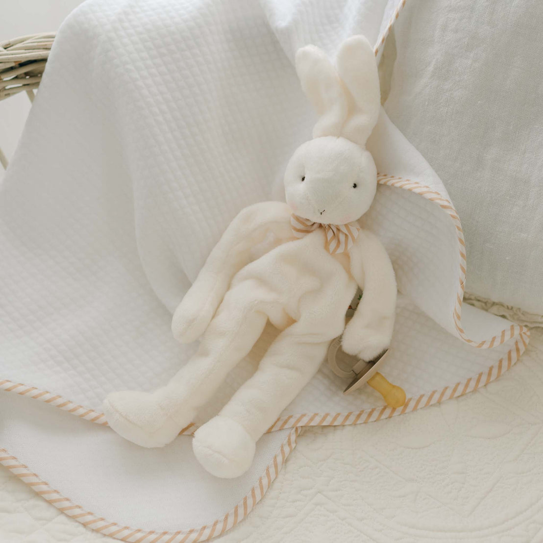 The Theodore Bunny Buddy, dressed in a striped bow tie, sits on a soft velour blanket with beige trim. This charming scene includes the blanket draped over the side of a bassinet and features a beige pacifier holder clipped near the plush rabbit—making it an adorable baby gift idea.