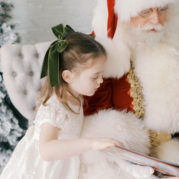 Young girl in a lace dress with a green bow sitting next to Santa Claus, holding a book.