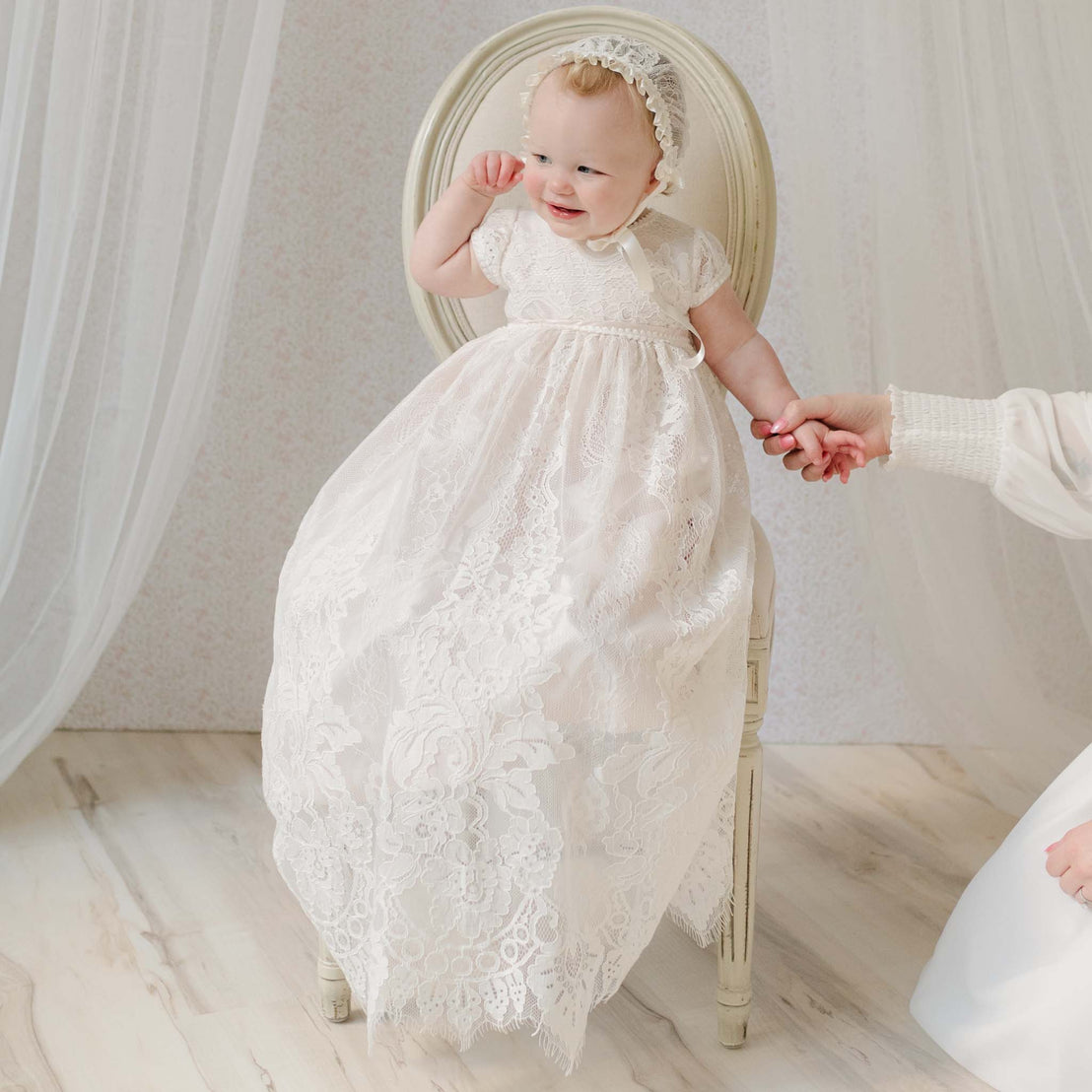 Baby in the Victoria Christening Gown & Bonnet sitting on a chair and holding hands with an adult off to the side. Part of the Baby Beau & Belle Christening Gowns Collection.