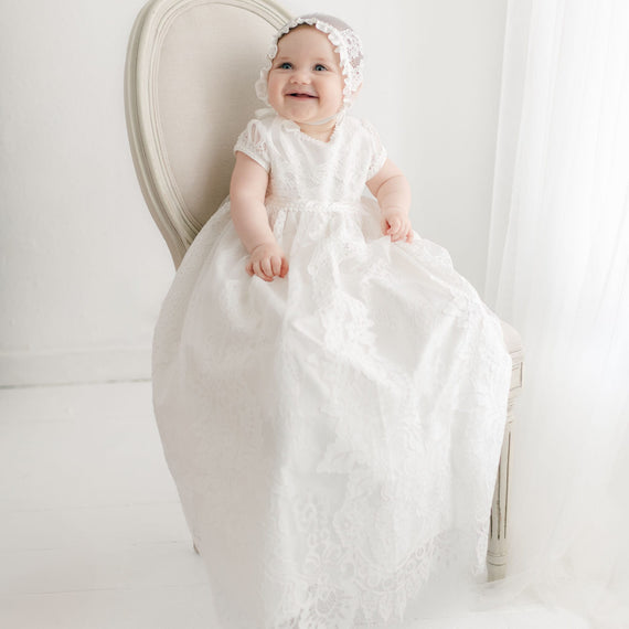 A smiling baby wearing a Victoria Puff Sleeve Christening Gown & Bonnet sits on a vintage, light-colored chair. The gown features ivory embroidered lace over a silk Dupioni lining. The The background is white and softly lit.