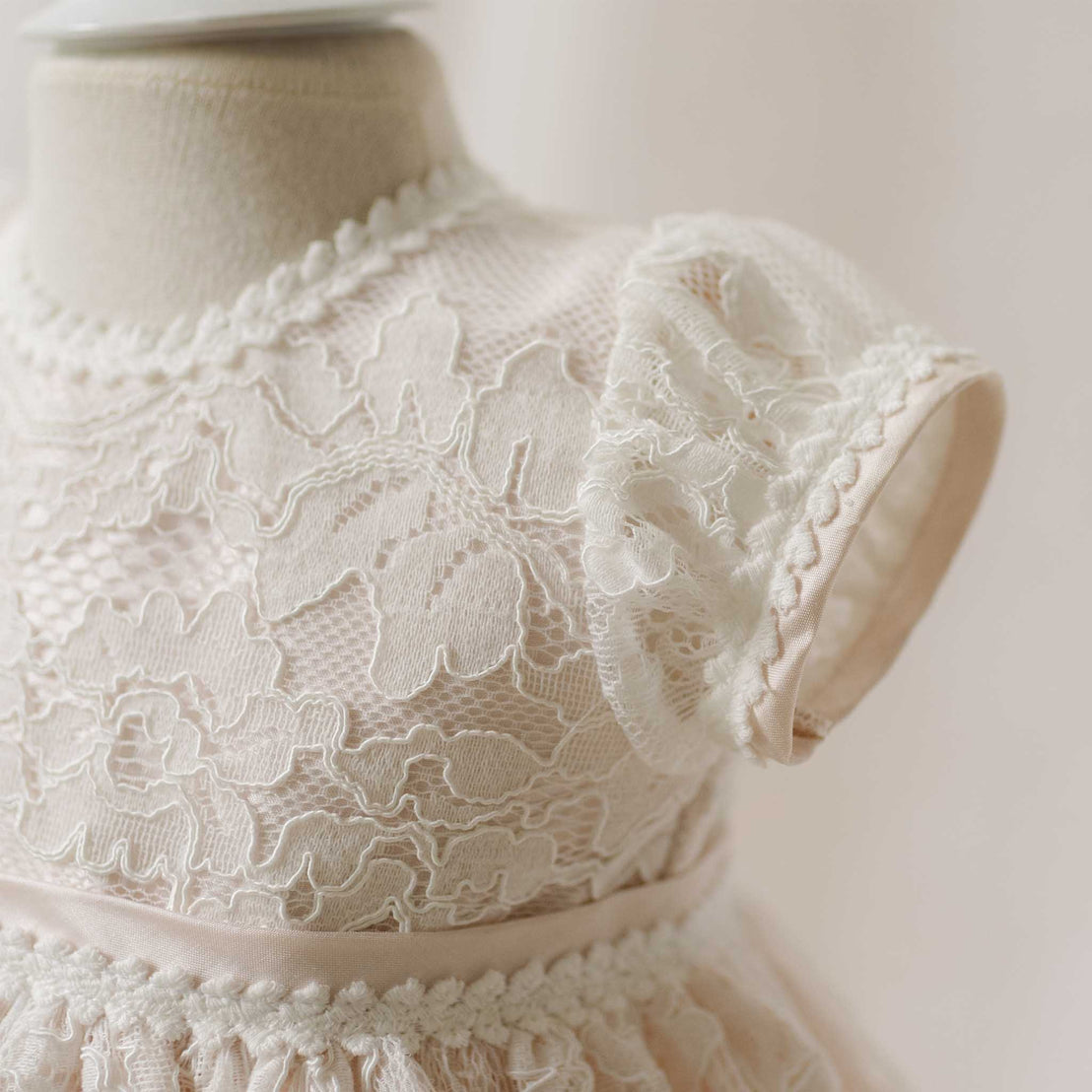 Close-up of the Victoria Christening Gown sleeve on a mannequin with a soft focus background. Details show the floral edge lace and silk trim. Part of the Baby Beau & Belle Christening Gowns Collection.