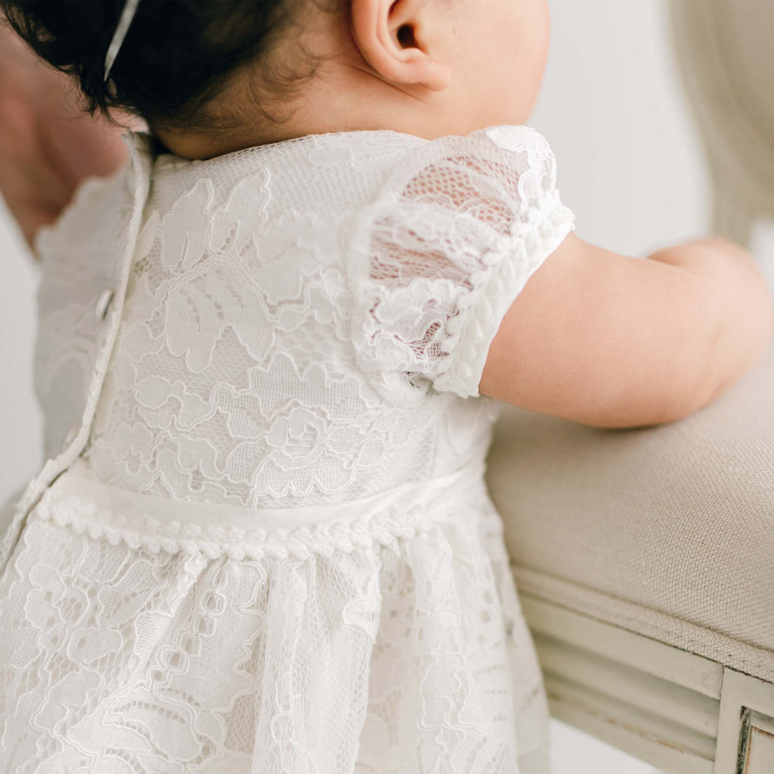 A baby in the Victoria Puff Sleeve Dress, crafted from white embroidered lace with intricate floral patterns, stands holding onto a cushioned chair. The baby's back is facing the camera, highlighting the dress's short, delicate sleeves and elegant button details made of silk Dupioni.