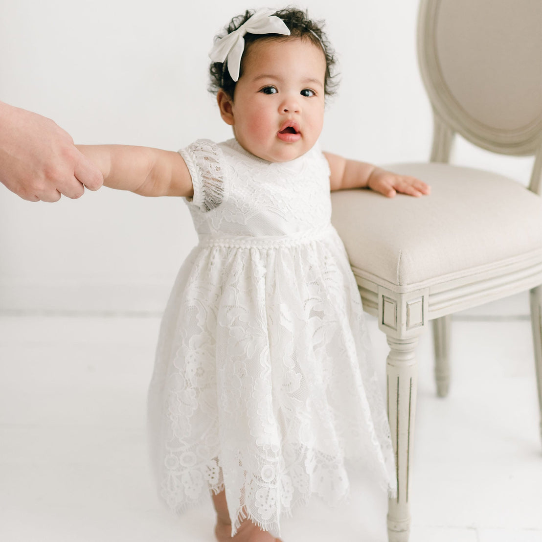 A toddler wearing the Victoria Puff Sleeve Dress and a matching headband stands holding onto a chair. An adult's hand gently supports her, guiding her as she balances in a bright room with light-colored furnishings.