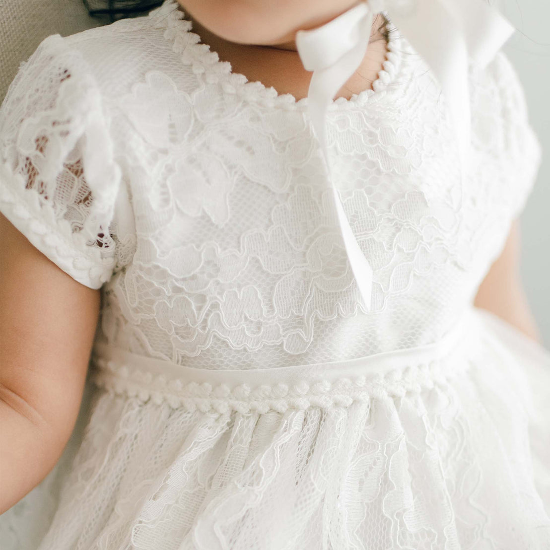 A baby wearing the exquisite Victoria Puff Sleeve Dress, crafted from embroidered lace showcasing intricate floral patterns and adorned with a satin ribbon around the waist. This charming dress features short sleeves and a subtle bow near the neckline, perfectly complementing the baby's partially visible face.