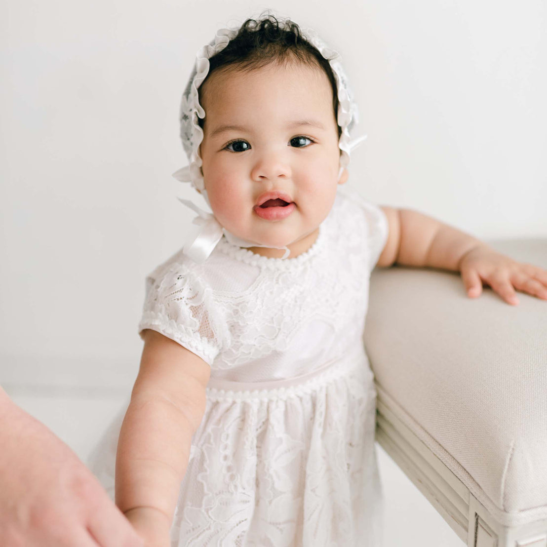 A baby in a silky Victoria Puff Sleeve dress adorned with embroidered lace stands while holding onto a beige cushioned surface. The little one has a curious expression and grips an adult’s hand for support, set against a soft, light background.
