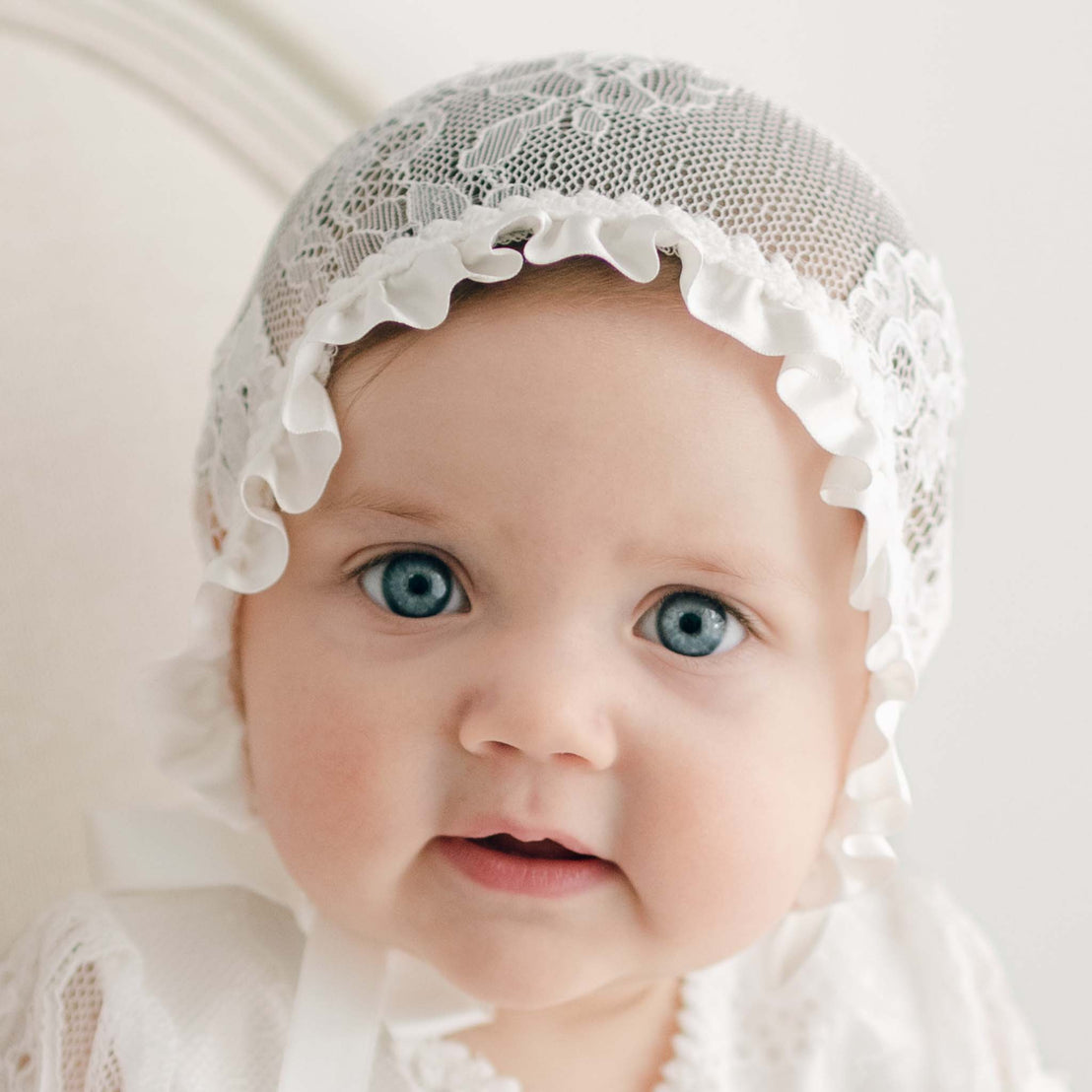 A close-up of a baby with blue eyes wearing a Victoria Lace Bonnet, looking directly at the camera with a gentle gaze.