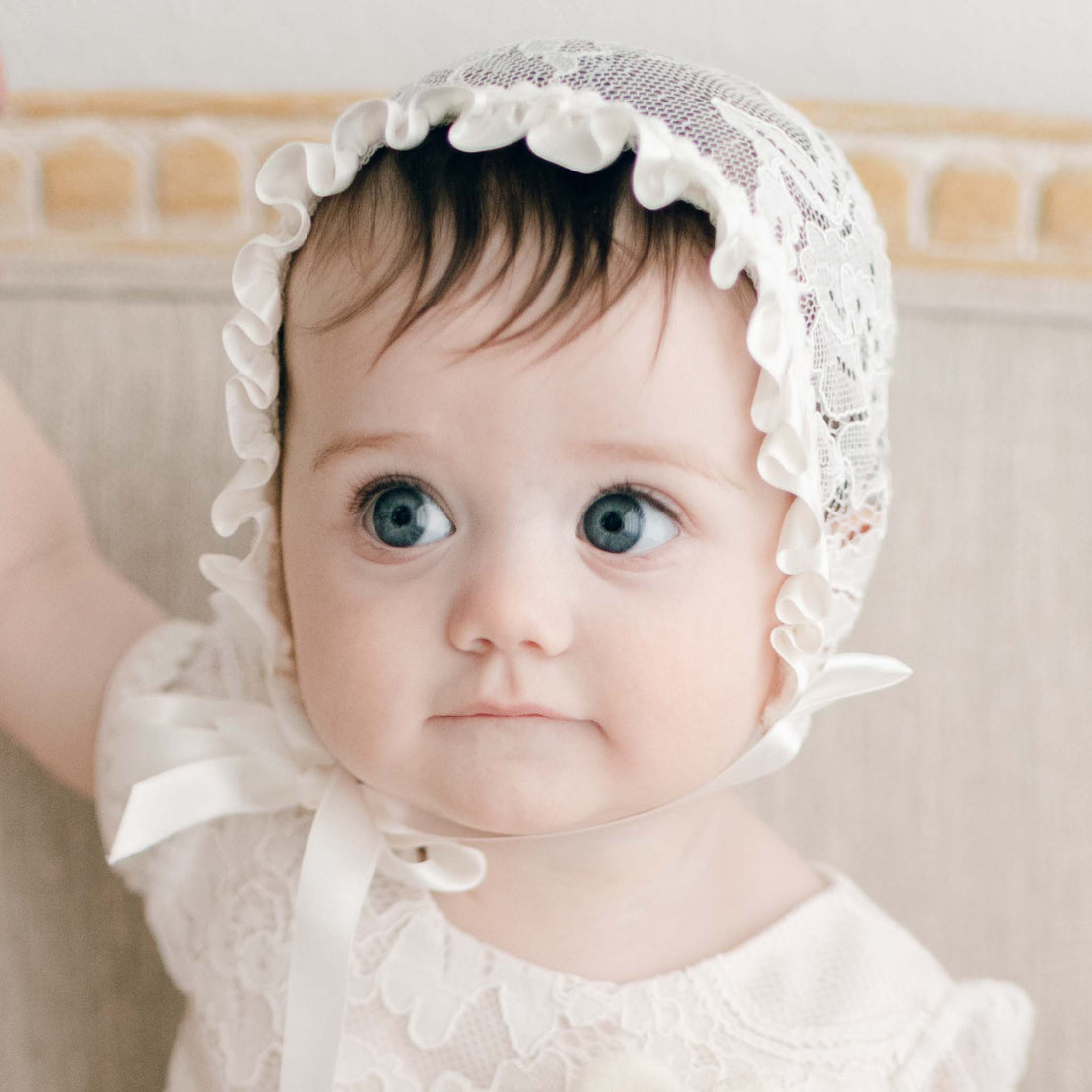 A baby girl with big blue eyes wearing the Victoria Lace Bonnet and matching dress looks to the side with slight smirk. The bonnet made with ivory floral lace and ivory silk ribbon is tied snug over her head.