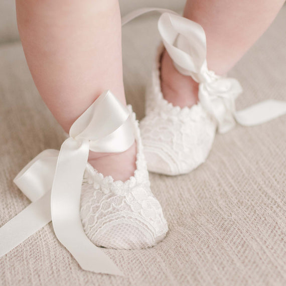 Close-up of a baby's feet wearing Victoria Lace Booties, tied with large silk ribbons, resting on a textured beige surface, perfectly complementing the christening gown.