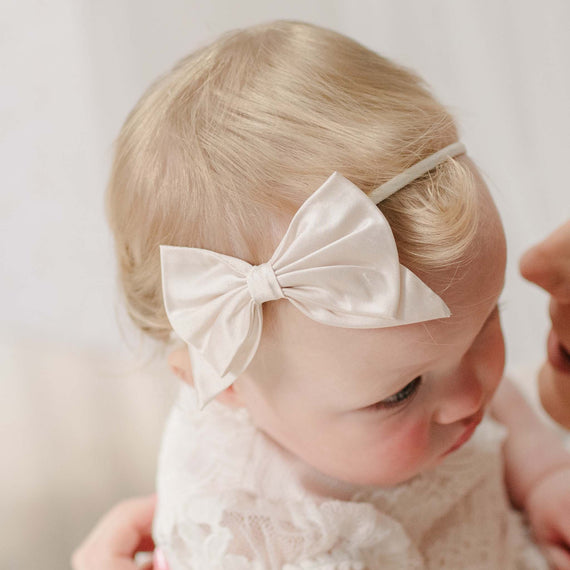 Baby wearing the Victoria Silk Bow Headband with a soft focus background. Detail shows a dupioni silk bow in pink champagne. Perfect for unique baby gifts.