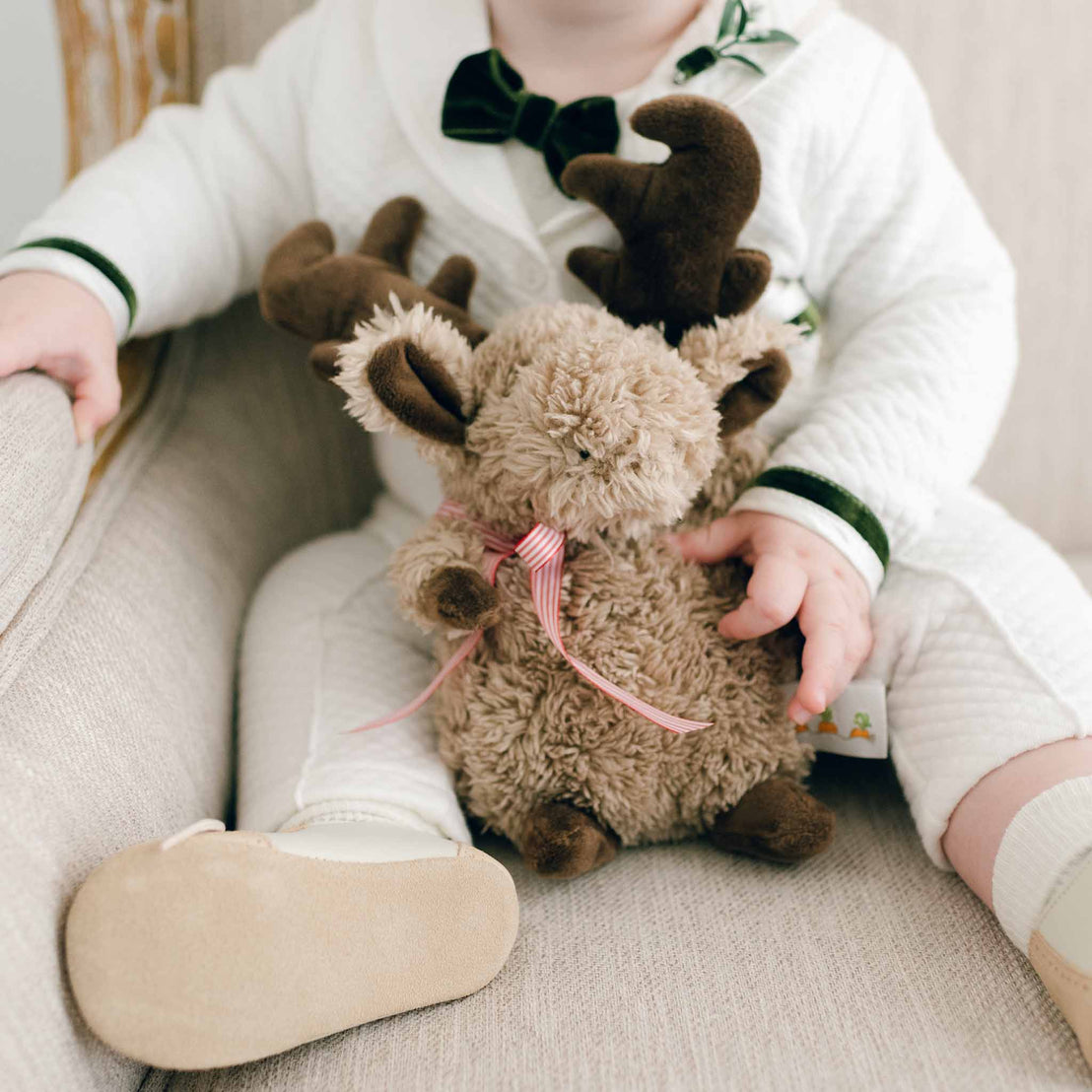 A baby-safe scene showcases a little one in a white onesie and beige shoes, seated on a cushioned chair. The child is holding the Wee Reindeer, which features soft fur and a red striped bow, while focusing on the toy and their hands.
