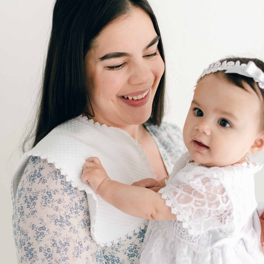 A woman with long dark hair smiles as she cradles a baby dressed in a white lace dress and headband. She is wearing a floral-patterned blouse and has the White Burp Cloth, made from quilted cotton, draped on her shoulder. Both exude warmth and joy against the soft white backdrop.