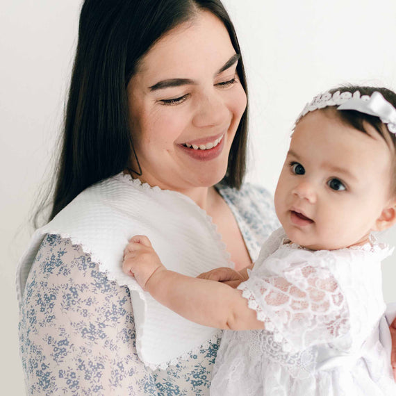 A woman with long dark hair smiles as she cradles a baby dressed in a white lace dress and headband. She is wearing a floral-patterned blouse and has the White Burp Cloth, made from quilted cotton, draped on her shoulder. Both exude warmth and joy against the soft white backdrop.