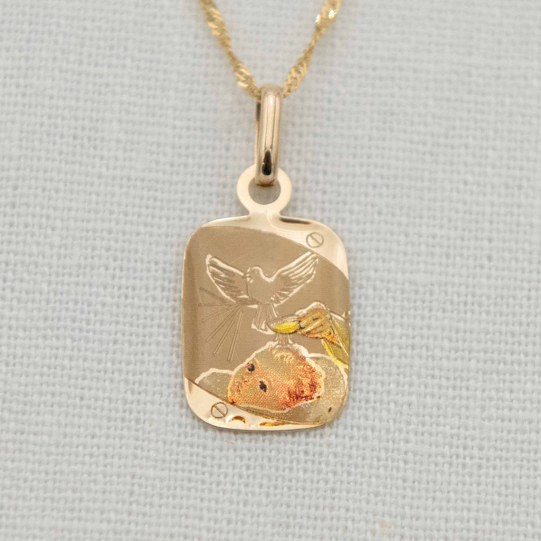 A 14k gold color etched blessing charm with a fine chain, depicting a dove with spread wings above a baby’s head as holy water is poured.