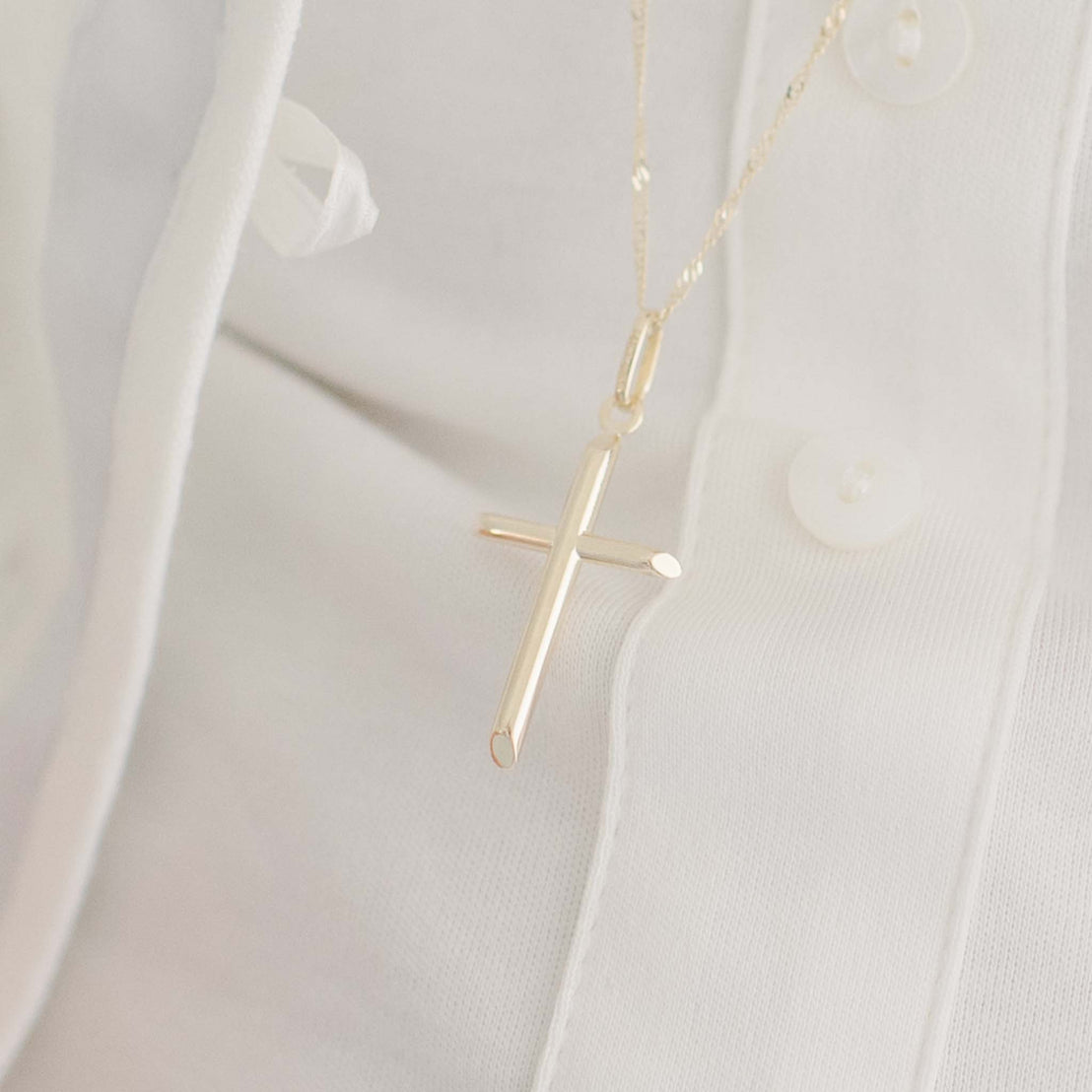 Close-up of a 14k Gold Cylindrical 'INRI' Cross Charm With Chain elegantly draped over a white button-up shirt. The soft, neutral background accentuates the simplicity and grace of this Christening jewelry.