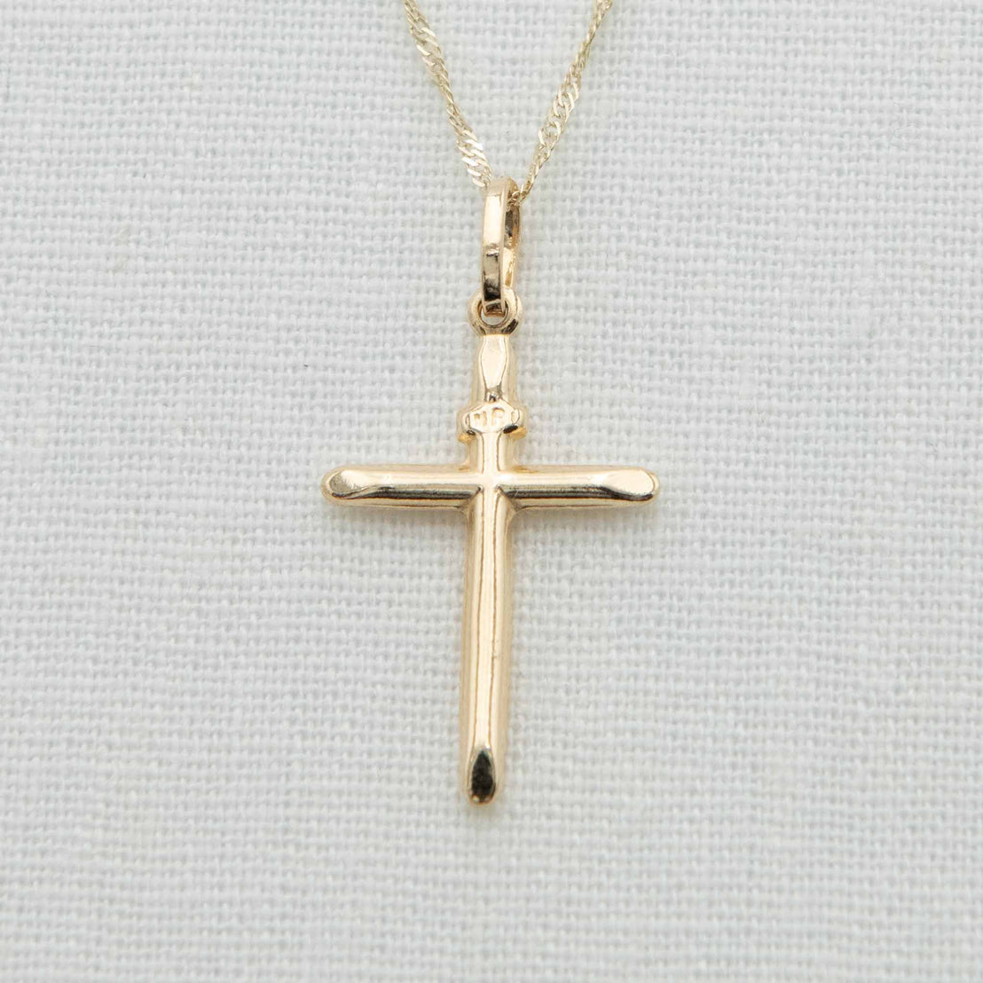 A 14k Gold Cylindrical 'INRI' Cross Charm with Chain, featuring a simple and elegant design, hangs delicately against a light fabric background. Its smooth edges and shiny finish make it ideal as Christening jewelry.
