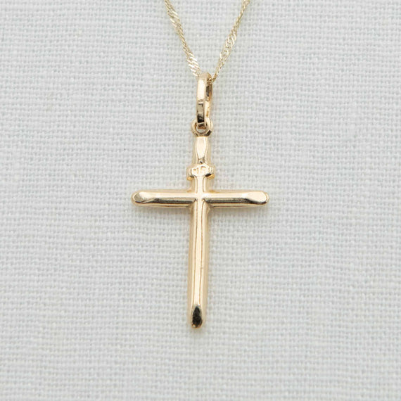 A 14k Gold Cylindrical 'INRI' Cross Charm with Chain, featuring a simple and elegant design, hangs delicately against a light fabric background. Its smooth edges and shiny finish make it ideal as Christening jewelry.