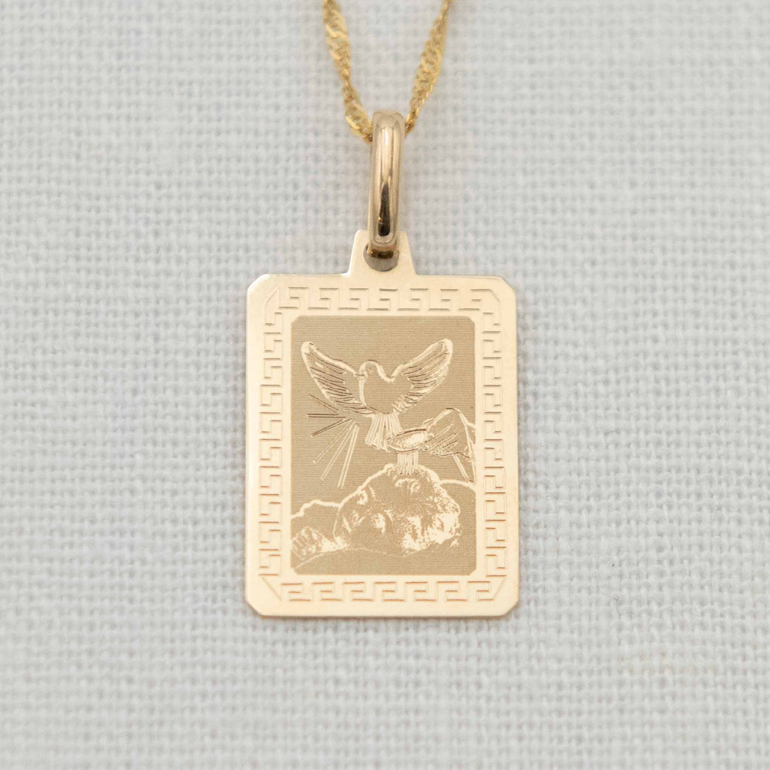 A 14k gold etched blessing charm with a fine chain, featuring a dove with open wings above a baby’s head as holy water is poured, framed by a decorative border.