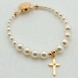 A pearl and gold bracelet featuring cream pearl beads, a gold clasp with a round medallion, and a dangling gold cross charm with a small clear stone, displayed on a textured white fabric background.