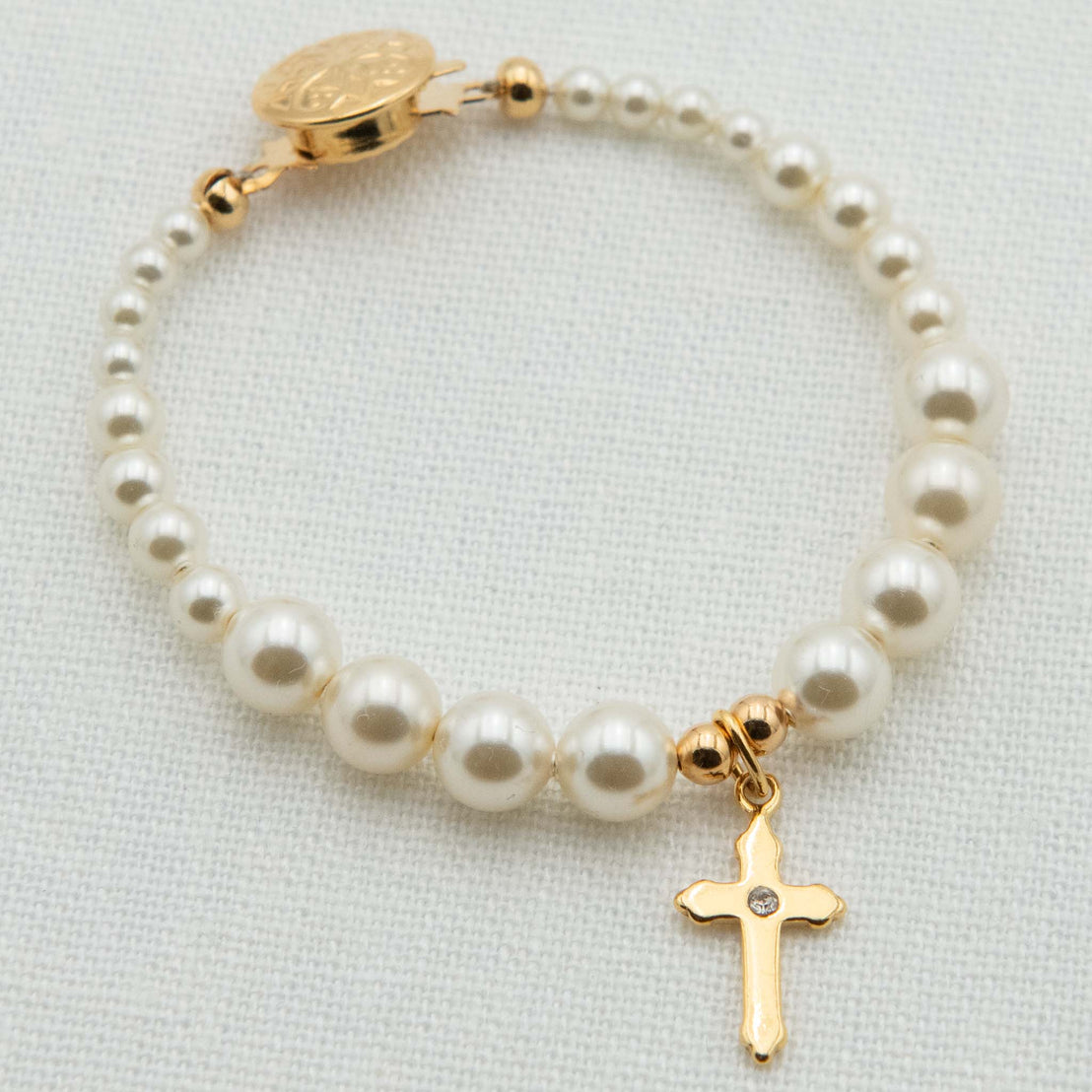 A pearl and gold bracelet featuring cream pearl beads, a gold clasp with a round medallion, and a dangling gold cross charm with a small clear stone, displayed on a textured white fabric background.