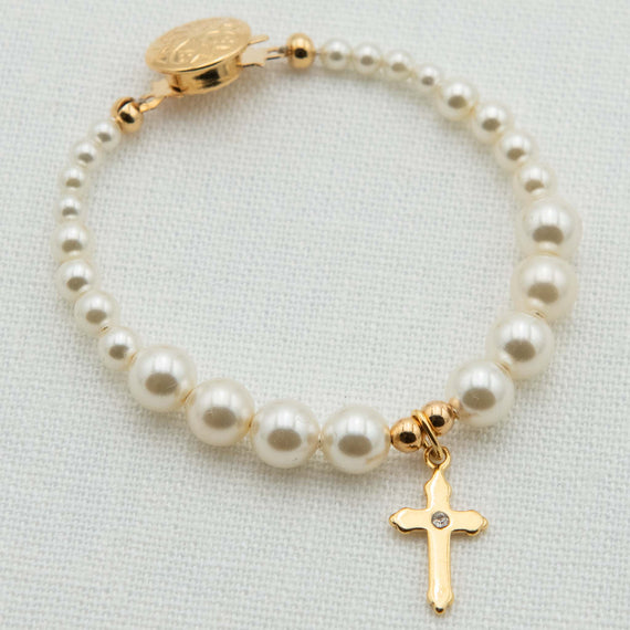 A pearl and gold bracelet featuring cream pearl beads, a gold clasp with a round medallion, and a dangling gold cross charm with a small clear stone, displayed on a textured white fabric background.