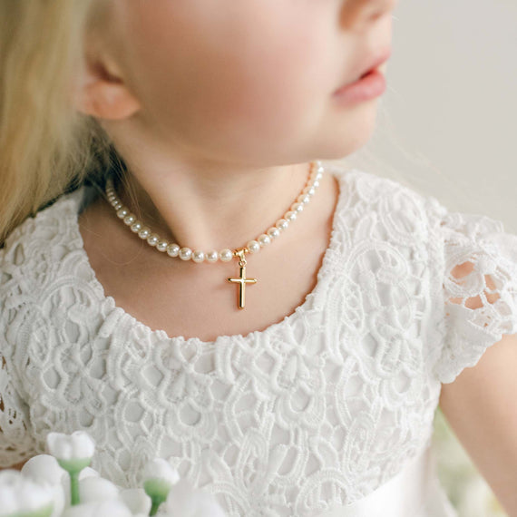 A close-up of a child wearing the Cream Luster Pearl Necklace with a small gold cross pendant, styled with a white lace dress.