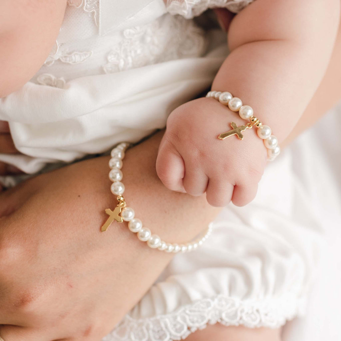 Close-up of a hand and child's hand holding together, both wearing pearl bracelets with gold crosses.