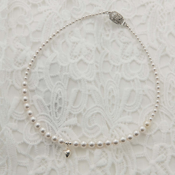 White pearl necklace featuring a small silver heart pendant, on a white lace backdrop.