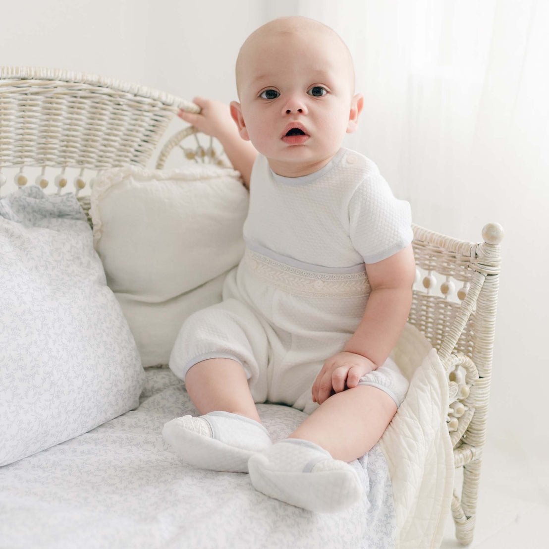 Baby seated on a vintage wicker crib adorned with soft cushions and blankets, wearing the Harrison Short Sleeve Romper with quilted fabric, lace trim, and pale blue accents.