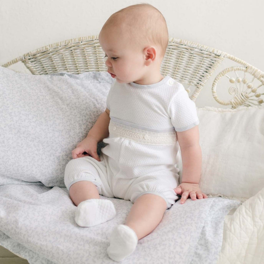 Baby sitting on a soft, patterned cushion in the Harrison Short Sleeve Romper, featuring quilted fabric with lace detailing and light blue accents.
