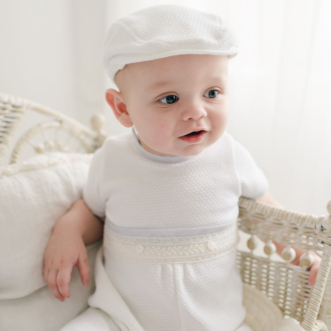 Baby wearing the Harrison Short Sleeve Romper with quilted fabric and lace detailing, paired with a matching white quilted newsboy cap, seated in a vintage wicker crib.