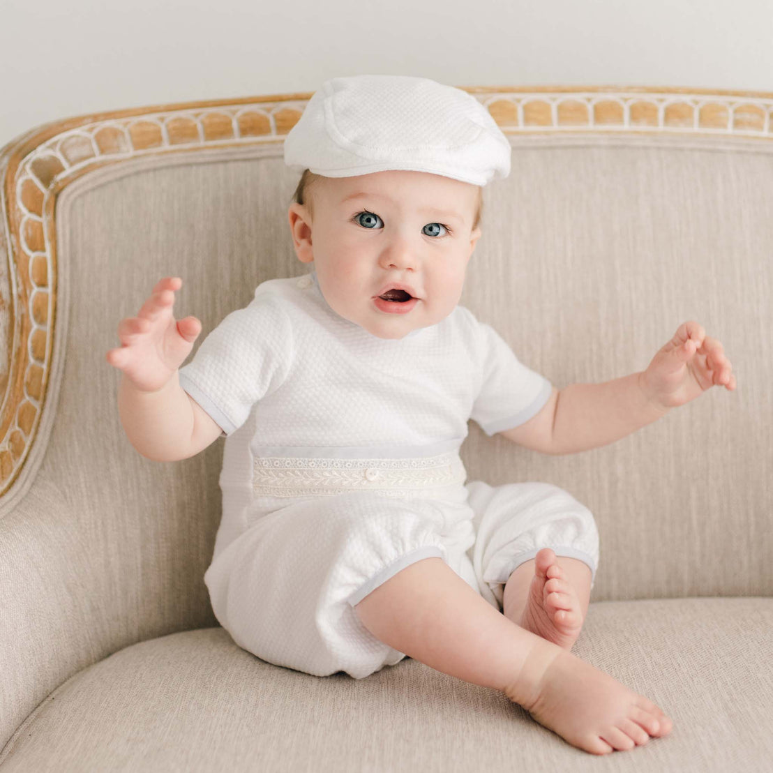 Baby wearing the Harrison Short Sleeve Romper, seated on a beige armchair, featuring the textured quilted fabric, lace waistband, and light blue accents, paired with a matching cap.