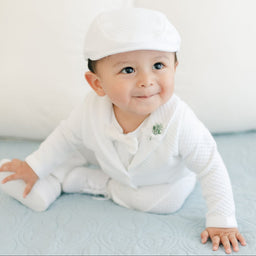A baby boy in the Elijah 3-Piece Suit and cap is sitting on a light blue blanket. The outfit, perfect for a special occasion, features textured cotton. He has a slight smile and is looking slightly upward, with white pillows in the background.