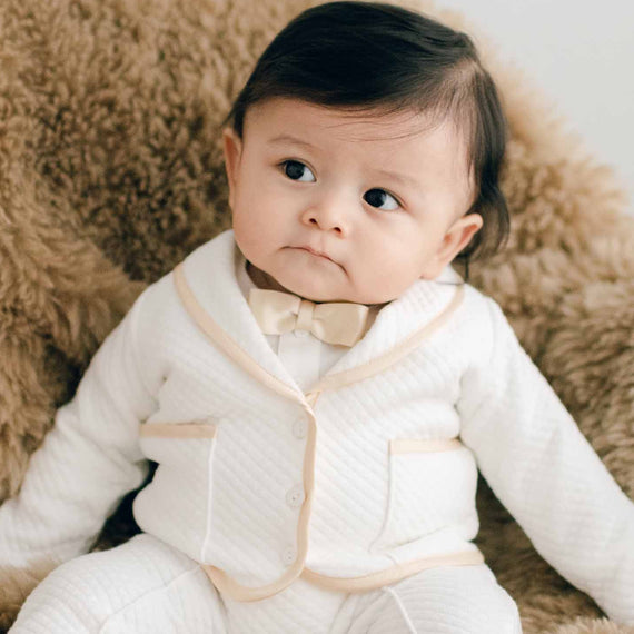 Baby sitting on a chair with brown fur. He is wearing the Liam 3-Piece Suit made with soft ivory quilted cotton and champagne silk trim. He is also wearing a champagne silk bowtie 