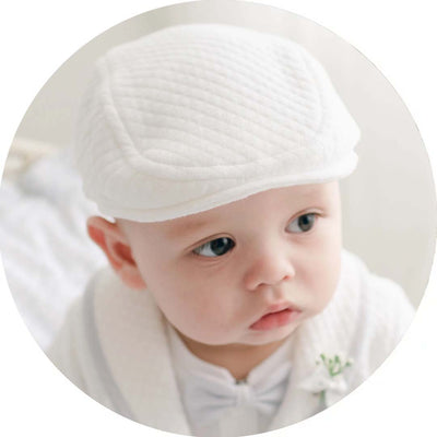 A baby wearing a textured white hat and matching outfit with a small boutonnière. The baby has a curious expression and is looking slightly to the side.