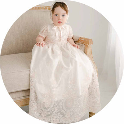 A baby wearing a white lace gown and bonnet sits on a vintage-style beige sofa. The setting is minimalistic with soft lighting.