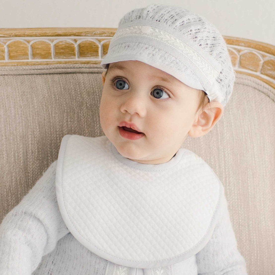 A baby boy wears the Harrison Bib made with a soft textured white cotton and features blue linen binding around the perimeter. He is also wearing the Harrison Knit Hat