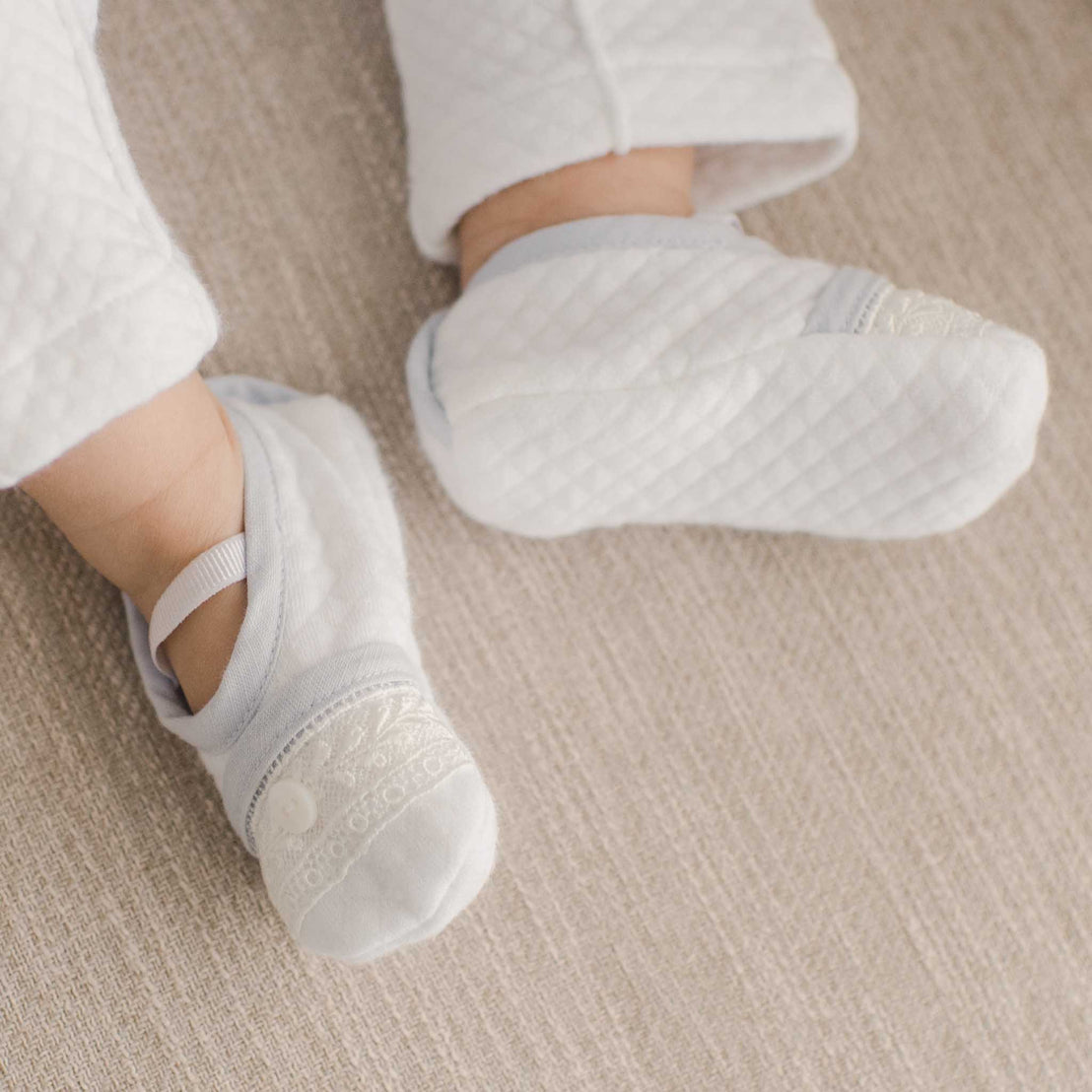 A baby's feet resting on a beige chair, wearing the Harrison Booties with quilted fabric and lace detailing for a cozy, stylish finish.