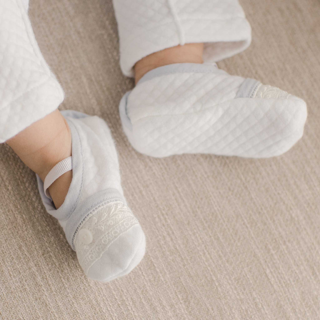 A baby wears a pair of Harrison Booties made with soft white textured cotton and a soft elastic straps with an ivory Venice lace, sky blue linen blend trim and a button on the toe 
