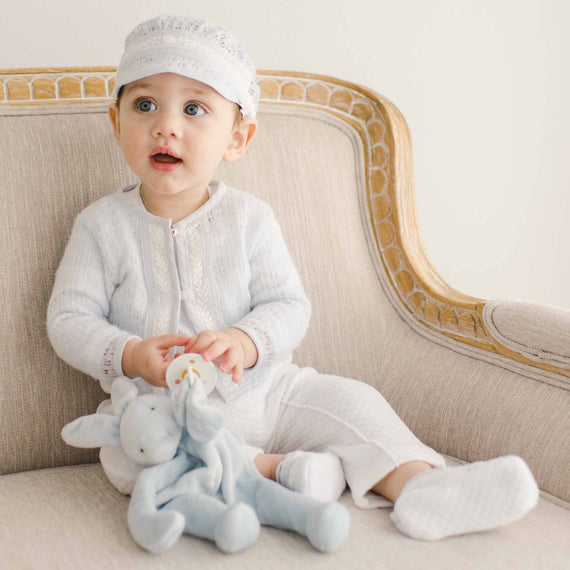 A baby sitting on a beige chair holding a plush blue bunny while dressed in the Harrison Sweater 3-Piece Pant Suit, including the knit sweater and lace-trimmed cap.