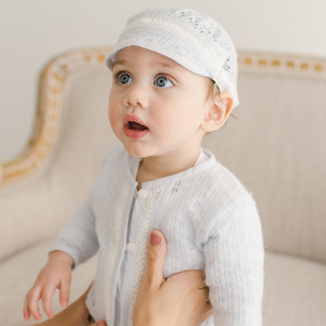 A baby wearing the Harrison Blue Knit Sweater with intricate lace detailing and white buttons, gently held by someone while seated on an upholstered chair.