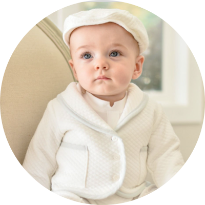 A baby wearing a white quilted outfit and a matching cap sits on a beige chair. The background is softly blurred, creating a serene and gentle atmosphere.